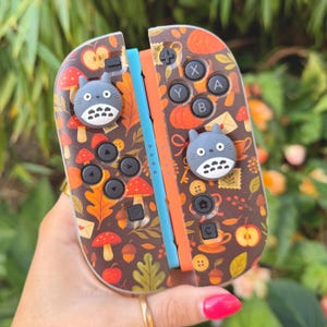 May include: Custom Nintendo Switch Joy-Con controllers with a brown autumn design featuring mushrooms, leaves, and apples. The controllers have a blue and orange accent and include two grey Totoro character thumb grips.