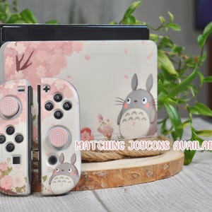 May include: A pink and white Nintendo Switch console with a Totoro design. The console has a pink and white floral design with a Totoro character on the front. The console is sitting on a wooden surface. Matching Joycons available.