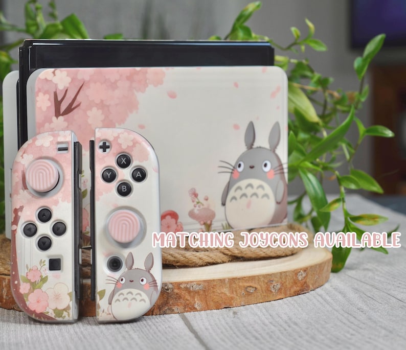 May include: A pink and white Nintendo Switch console with a Totoro design. The console has a pink and white floral design with a Totoro character on the front. The console is sitting on a wooden surface. Matching Joycons available.