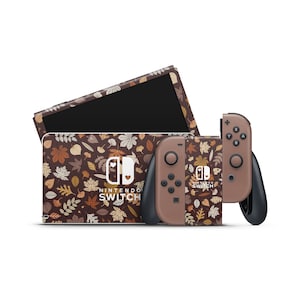 Op de afbeelding: Een Nintendo Switch-console en twee controllers met een bruin en beige herfstbladerenpatroon. De console heeft de tekst "Nintendo Switch" en een klein hartpictogram. De controllers hebben de tekst "Nintendo Switch" en een klein hartpictogram.