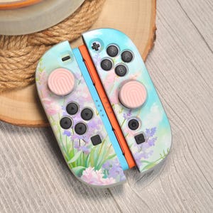 May include: A pair of pastel blue and orange game controllers with a floral design. The controllers have pink thumbsticks and black buttons. The design includes lavender and other flowers.