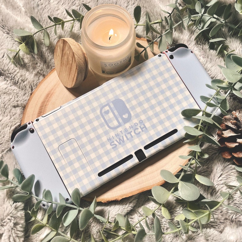 May include: A white Nintendo Switch console with a light blue and white gingham pattern and the text "Nintendo Switch" on the front. The console is sitting on a wooden surface with a jar of candle and a wooden lid.