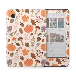 Może przedstawiać: Czytnik e-booków Kindle w kolorze kremowym z etui w jesiennym stylu. Etui ma wzór pomarańczowych dyni, brązowych żołędzi, czerwonych jagód i kolorowych liści na kremowym tle.