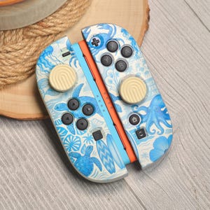 Peut inclure: Une paire de manettes Joy-Con Nintendo Switch bleues et blanches. Les manettes présentent un motif de vie marine avec des tortues, des pieuvres et des planches de surf. Les boutons sont noirs et les joysticks beiges.