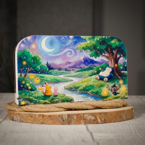 Op de afbeelding: Een kleurrijke illustratie van een serene landschap met een rivier, bergen en een halve maan in een sterrenhemel. Verschillende stripfiguren zijn afgebeeld in de scène, waaronder een geel wezen en een blauw, slapend personage onder een boom. Het kunstwerk wordt getoond op een houten basis.