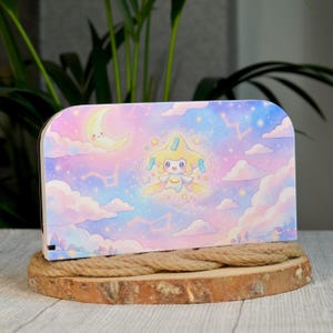Può includere: Una cover rettangolare per dock di console con un'illustrazione di un cielo dai toni pastello, un personaggio centrale giallo a forma di stella, una luna crescente sorridente, nuvole rosa e blu e costellazioni luminose.