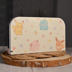 Może przedstawiać: Kremowe etui na Nintendo Switch z postaciami z kreskówek Pikachu, Bulbasaur, Jigglypuff i Eevee. Etui jest ozdobione kolorowymi gwiazdkami, kwiatami i truskawkami. Etui stoi na drewnianym plastrze.