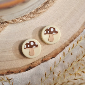 May include: Two white round buttons with brown and white mushroom designs. The mushrooms have white dots on the top and a brown stem.