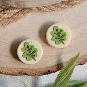 以下が含まれることがあります： 緑の葉のデザインが施された、クリーム色の丸いボタンが2つ。ボタンは木製の表面に置かれています。