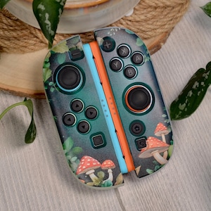 Może przedstawiać: Para kontrolerów Joy-Con do Nintendo Switch z niestandardowym wzorem. Kontrolery mają ciemnozielone tło z wzorem grzybów i liści. Przyciski są czarne, a boki niebieskie i pomarańczowe.