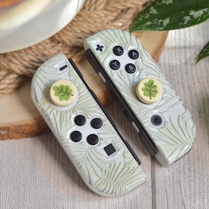 Peut inclure: Une paire de manettes Joy-Con blanches pour Nintendo Switch avec un motif de feuilles vertes et des boutons beiges. Les manettes ont des capuchons de boutons personnalisés avec des motifs de feuilles vertes.