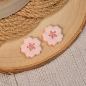 May include: Two light pink, five-petaled flower-shaped beads, each featuring a darker pink, branched stamen design in the center. These small, flat-backed floral embellishments are crafted with a smooth, matte finish and a symmetrical shape.