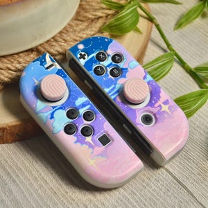 Puede incluir: Un par de mandos Nintendo Switch Joy-Con rosa y azul con diseños personalizados que incluyen nubes, estrellas y lunas. Los mandos tienen botones rosa y azul y palancas analógicas rosas.