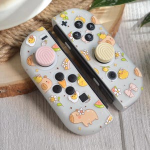 May include: Two gray Nintendo Switch Joy-Con controllers with a repeating pattern of cartoon animals, flowers, and fruit. The controllers have black buttons and pink and beige thumbsticks. The design includes a bear, chick, and capybara.