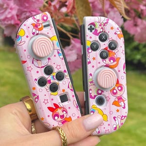 Puede incluir: Mandos Joy-Con de Nintendo Switch rosas con un diseño personalizado que presenta a los personajes de las Chicas Superpoderosas. Los mandos tienen botones rosas y un fondo rosa y blanco con estrellas y arcoíris. Los personajes están en varias poses y tienen contornos rosas, blancos y negros.