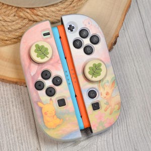 May include: A pair of Nintendo Switch Joy-Con controllers with custom artwork. The controllers feature a pastel watercolor design with Pikachu and Leafeon illustrations. The thumbstick caps have a green leaf design. The controllers are orange, blue, and silver.