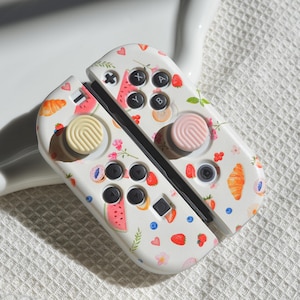 May include: White Nintendo Switch Joy-Con controllers with a custom design featuring a watermelon, strawberries, croissants, and flowers. The controllers have pink and cream colored button caps.