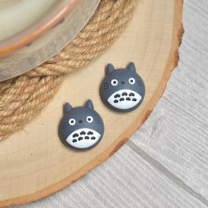 Op de afbeelding: Twee grijze Totoro-vormige decoraties met witte en zwarte details. Ze liggen op een houten schijf, in de buurt van een touwachtig object. De items hebben een cartoonachtig uiterlijk, met ronde ogen en een lachende mond. De achtergrond is een lichtgekleurd houten oppervlak.