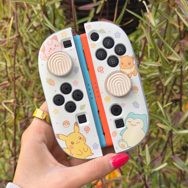 May include: A pair of white game controllers featuring colorful illustrations of Pikachu, Eevee, Snorlax, and Jigglypuff, accented with small flowers, stars, and berries. Each controller has a beige thumbstick cover with a textured, circular pattern.