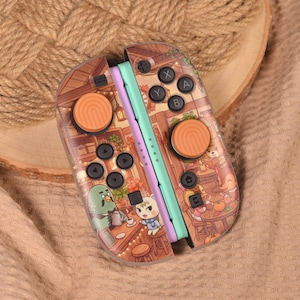 Puede incluir: Un par de mandos de juego con una funda personalizada que muestra una escena de cafetería. Los mandos tienen rieles laterales en verde claro y lavanda, botones negros y dos cubiertas de joystick naranjas con textura.