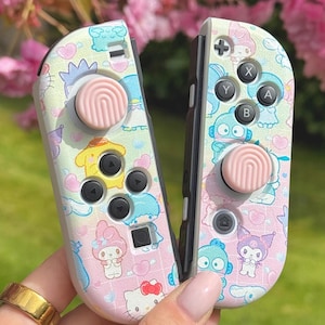 May include: A pair of pink and white Nintendo Switch Joy-Con controllers with custom Hello Kitty and Sanrio character designs. The controllers feature pink and white buttons and pink analog sticks.