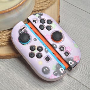 Peut inclure: Une paire de manettes Joy-Con Nintendo Switch lavande. Les manettes présentent un motif répété de créatures noires et duveteuses et d'étoiles colorées. Les boutons sont noirs et les rails sont orange et bleu.