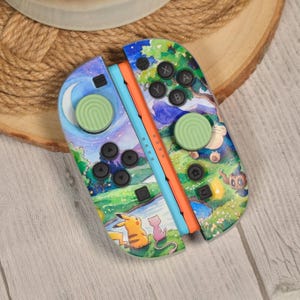 Op de afbeelding: Aangepaste Nintendo Switch Joy-Con controllers. De controllers hebben een kleurrijk aquarel landschapsontwerp met stripfiguren, waaronder Pikachu. De knoppen zijn zwart en de joysticks lichtgroen.
