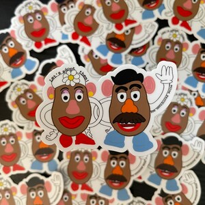 May include: A pile of white stickers featuring cartoon characters of Mr. and Mrs. Potato Head. The characters are brown and have white eyes, red lips, and black eyebrows. The text on the sticker reads "Let's adopt them!"