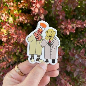 May include: A sticker featuring two Muppet characters, one red with orange hair and the other yellow with a mustache, both wearing white lab coats. The red Muppet is holding a heart in its hand.