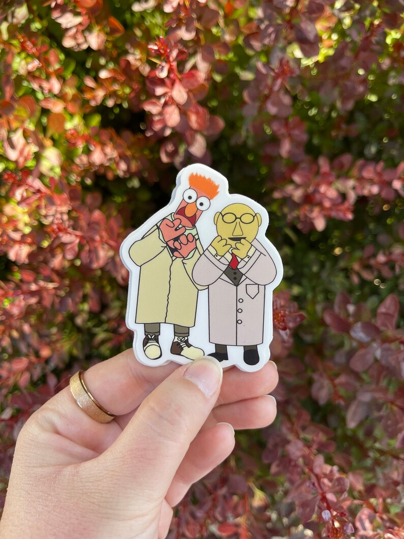 May include: A sticker featuring two Muppet characters, one red with orange hair and the other yellow with a mustache, both wearing white lab coats. The red Muppet is holding a heart in its hand.