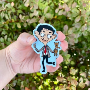 May include: A cartoon sticker of Mr. Bean, a character from the British sitcom, Mr. Bean. He is wearing a gray suit, red tie, and brown shoes. He is holding a brown teddy bear in his right hand and waving with his left hand. The sticker is on a light blue background.