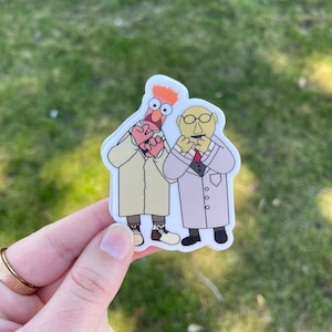 May include: A sticker featuring two Muppet characters, one with red hair and a white lab coat, the other with a yellow face and a white lab coat. Both characters are holding their hands to their mouths.