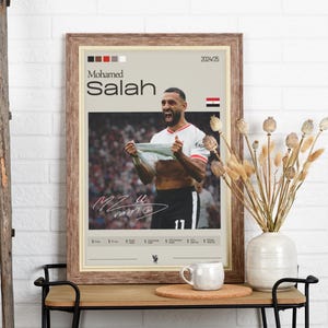 Op de afbeelding: Een ingelijste poster met een foto van Mohamed Salah, een voetballer, die een doelpunt viert. De poster heeft een bruine houten lijst en een witte achtergrond met de tekst "Mohamed Salah" en "2024/25" in zwart. De poster bevat ook een kleine Egyptische vlag.