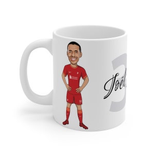Könnte beinhalten: Weiße Keramik-Tasse mit einer Cartoon-Illustration eines Fußballspielers in roter Kleidung. Die Tasse trägt den Namen des Spielers und die Nummer 32. Die Tasse hat einen weißen Henkel und ein Fassungsvermögen von ca. 325 ml.