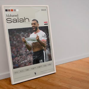 Op de afbeelding: Een witte geframed poster met een foto van Mohamed Salah die een doelpunt viert. De poster heeft een witte achtergrond met zwarte tekst die "Mohamed Salah" en "2024/25" zegt. De poster heeft ook een kleine Egyptische vlag in de rechterbovenhoek.