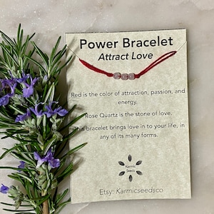 May include: Red string bracelet with three rose quartz beads. The card reads "Power Bracelet Attract Love" and includes information about the meaning of the color red and rose quartz. The card also includes the Etsy shop name "Karmicseedsco".