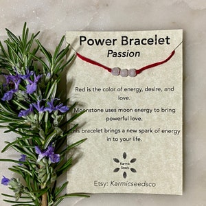 Puede incluir: Un brazalete de cuerda roja con tres cuentas de piedra lunar. La tarjeta dice "Power Bracelet Passion" e incluye el texto "El rojo es el color de la energía, el deseo y el amor. La piedra lunar utiliza la energía lunar para traer un amor poderoso. Este brazalete trae una nueva chispa de energía a tu vida." La tarjeta también incluye el texto "Karmic Seeds" y "Etsy: Karmicseedsco".