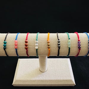 May include: Ten bracelets with different colored cords and two gemstone beads each. The bracelets are displayed on a beige fabric stand.