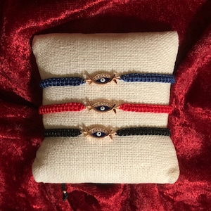 May include: Three string bracelets with a gold evil eye charm. The bracelets are red, blue, and black.