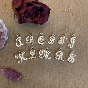 May include: Silver charm necklace pendants in the shape of the letters A through S. The charms are arranged in two rows on a wooden surface.
