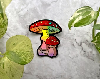 May include: A holographic sticker of two mushrooms. The larger mushroom has a red and white cap with a rainbow iridescent sheen. The smaller mushroom is red with white spots. The mushrooms are on a white marble background.