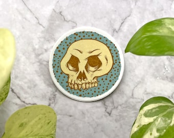 May include: Circular sticker with a skull illustration. The skull is beige with brown details, set against a teal background with brown dots. The sticker has a white border and is likely for decorative purposes.