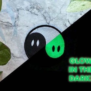 Op de afbeelding: Een zwart-wit yin yang symbool met een groene helft die in het donker gloeit. De tekst "GLOW IN THE DARK!" staat onder het symbool.