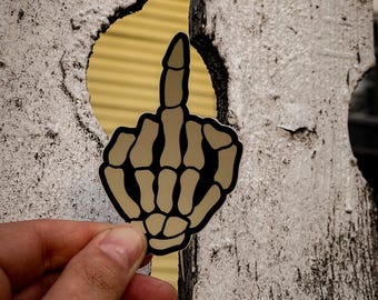 May include: A sticker of a skeleton hand giving the middle finger, in a beige colour with black outlines, on a white wooden background.