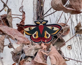 May include: A red, yellow and black moth sticker with large eyes, attached to a weathered wooden surface with dried brown leaves.