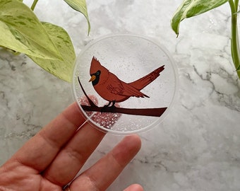 May include: A clear round sticker featuring a red cardinal perched on a branch. The sticker has a snowy background with a white border.