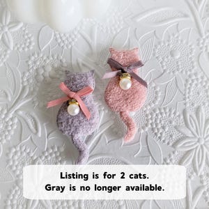 May include: Two cat-shaped brooches. One is lavender, the other is pink. Each has a ribbon bow, a faux pearl, and a gold-colored accent. The text on the image says "Listing is for 2 cats. Gray is no longer available."