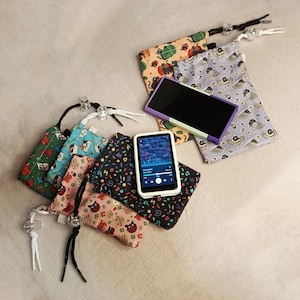 May include: A selection of small drawstring pouches with various patterns, such as cats, dogs, and abstract designs. An MP3 player with a white case and a purple-framed tablet are also visible, alongside the pouches.