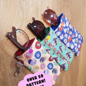 May include: Three different patterned glasses cases with a key chain attached to one of them. The cases are made of fabric and have a variety of colorful designs. The text "OVER 50 PATTERN! (PT.2)" is written on a pink cloud.