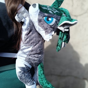 Op de afbeelding: Een grijze en groene pluche wolf rugzak met een blauw oog. De wolf draagt een groene fluwelen cape.
